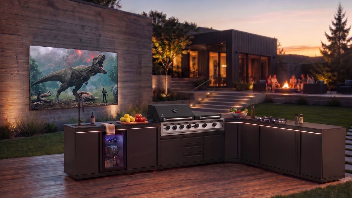 A modern outdoor kitchen setup on a wooden deck at dusk, featuring a built-in gas grill, stainless steel appliances, and dark cabinetry. A large wall-mounted screen displays a dinosaur scene, and a patio area with warm lighting and people is visible in the background.